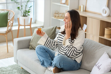 Young woman with glass of Turkish tea talking by mobile phone on sofa at home
