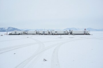 Obraz premium An oil depot on a snowy plain, with snow-covered tanks and pipelines. The background shows a gray sky and distant snowy mountains