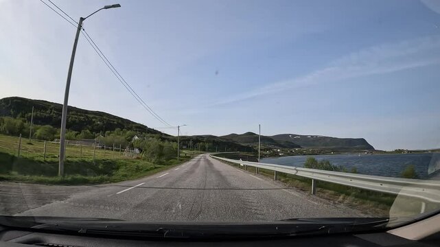 Driving along Norway's northern tundra on E69 highway. The sea lies to the right, while sparse forest, rocks, small meadows, and a few houses line the left. A peaceful coastal journey through nature.