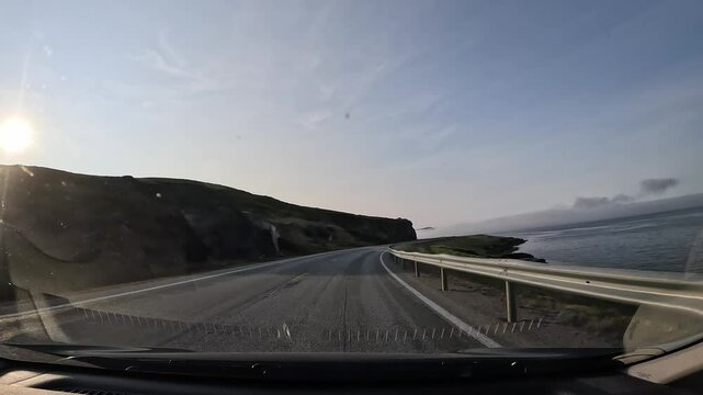 Driving along Norway's E69 road in the north. Rugged rocky cliffs on the right, the sea on the left. A band of misty clouds hovers above the water, with the low sun glowing in the distance.