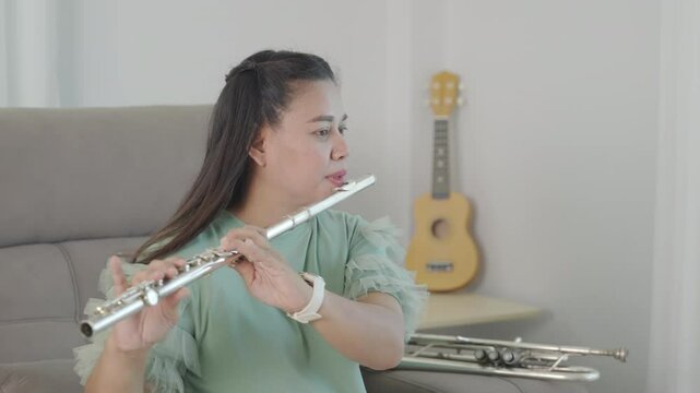 A woman in a green top, seated on a brown couch at home, blows into her silver flute with grace and skill. The living room ambiance adds a personal touch to her musical artistry.