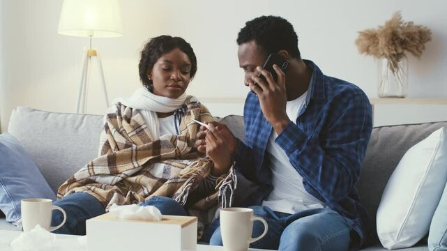 Two young adults, wrapped in blankets and looking uneasy, sit on a couch in their home. One is on the phone, likely seeking help while the other appears unwell. Tissues are nearby.