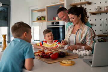 Caucasian family with two children preparing fresh ingredients in modern kitchen while following recipe on laptop during cooking lesson at home.