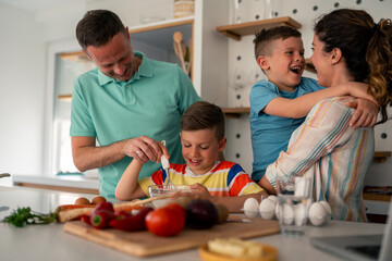Caucasian parents and children collaborate in modern kitchen, following digital recipe. Father wearing polo shirt guides cooking activity while mother shares joyful moment.