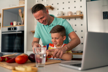 Caucasian father in mint polo shirt assists young son with food preparation in contemporary home kitchen while following recipe on nearby laptop computer.
