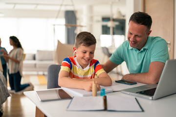Caucasian father in polo shirt guides young boy wearing colorful striped shirt with homework assignment using laptop and papers at home desk.