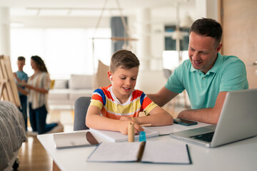 Caucasian father in mint polo shirt guides young boy wearing colorful striped shirt with homework assignment at home office desk with laptop.