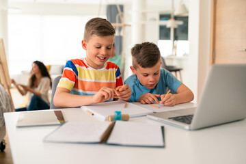 Obraz premium Caucasian elementary school brothers concentrate on homework assignments using laptop and notebooks at bright home study space while parent supervises.