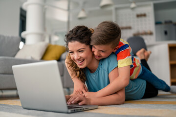Caucasian mother and young son in casual attire share laptop screen while relaxing on carpeted living room floor, displaying genuine joy.