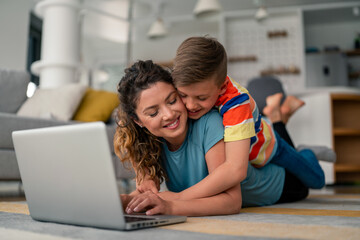 Caucasian mother and young son lie comfortably on living room carpet, sharing moments while using...