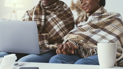 Two African American millennials sit closely together under blankets while working on a laptop. They are showing signs of illness while staying warm at home during recovery from flu or cold symptoms. - Powered by Adobe