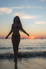 A woman in a patterned bikini stands at the water's edge during sunset, with sailboats silhouetted on the horizon and gentle waves lapping at her feet.