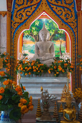 Serene white Buddha statue in meditative pose, surrounded by orange flowers, golden details, and a colorful mural in a traditional Thai temple.