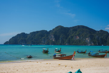Turquoise waters, limestone cliffs, and anchored longtail boats with colorful ribbons on a sandy beach under a clear blue sky in Koh Phi Phi, Thailand.