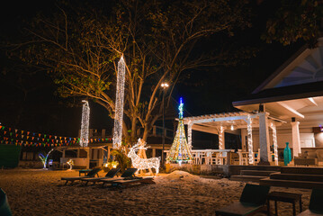Nighttime beach setting in Phuket, Thailand, featuring a decorated tree, illuminated reindeer, string lights, bar seating, and lounge chairs on the sand.