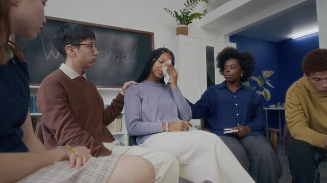 Medium wide shot of crying Black teen girl surrounded by empathic young Black female psychologist and multiethnic peers trying to support and help her to cope with crisis, sitting in chairs in class