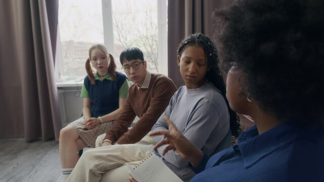 Side view shot of young African American psychologist speaking to diverse zoomers sitting in chairs in circle while leading group therapy session in modern class