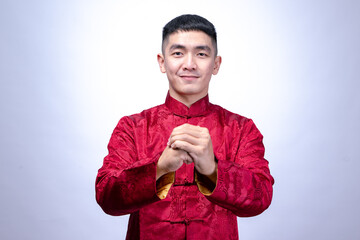 A smiling young man in a red traditional Chinese Tang suit performs a Gong Xi greeting gesture,...