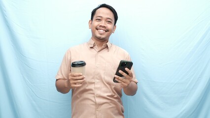 Fototapeta premium Smiling Asian man in a beige shirt holding a coffee cup in one hand and a smartphone in the other, standing against a light blue background.