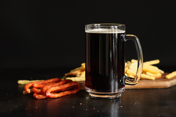 Glass with dark beer and snacks on dark background. Beer, French fries and hunter sausages. Side view of glass on wooden board and snacks. Dark beer and bread croutons. Traditional glass beer mug