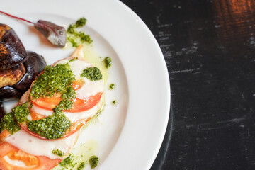 Caprese chicken with pesto and grilled eggplant on white plate over dark background.