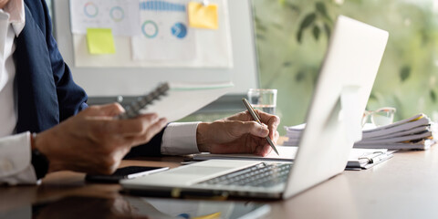 Elderly businessman taking notes while analyzing data on a laptop in a modern office