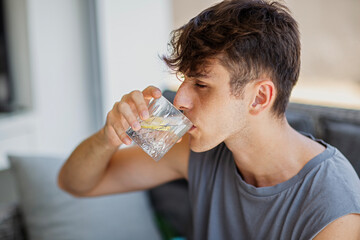 Young man drinking water with lemon from a glass on the sofa at home close-up