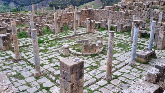 Revealing shot of the ancient Cosinus market, framed by impressive Roman columns at the historic site of Djemila, Algeria, with green hills and an overcast sky in the background