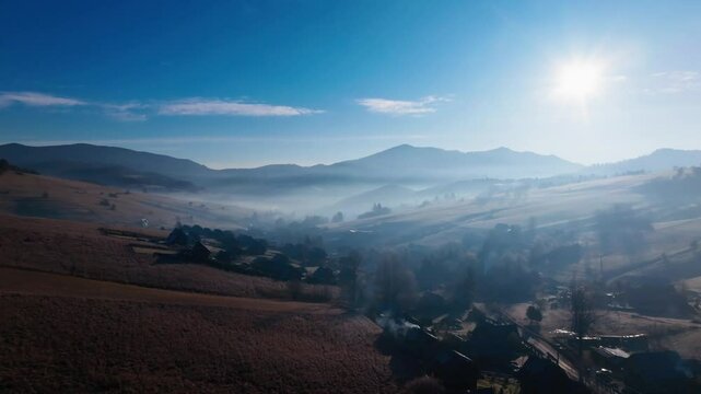 A serene aerial video of sunlight piercing through misty hills, revealing the natural charm of a tranquil valley and scattered trees in a rural winter landscape.