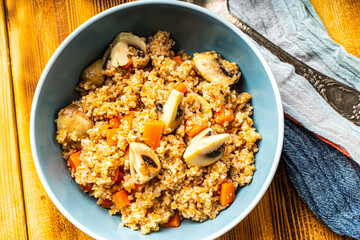 Boiled spelled or spelled porridge with mushrooms, carrots and spinach in a blue bowl on a wooden background with a napkin and spoon. Healthy eating concept. Vegetarian dish.