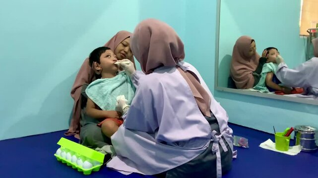 A child with cerebral palsy accompanied by his mother is undergoing oral stimulation using a special needs brush assisted by a speech therapist in the children's medical rehabilitation room.