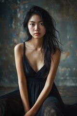 Fototapeta premium A striking image of an Asian woman in a black dress, sitting against a textured background with an intense gaze.