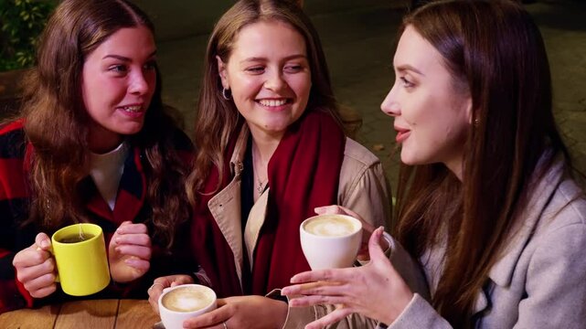 Three young women smiling and chatting while holding cups of coffee at an outdoor cafe during the evening.