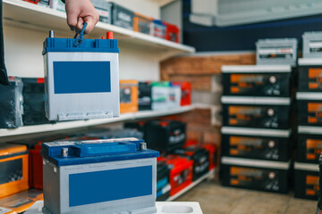 A person holding an automotive battery by its blue handle above another battery on a white surface. Shelves with car batteries and tools visible in the background