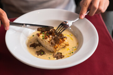 Hands of young man with knife and fork cutting fish steak and cream sauce garnished with slices of truffle on white plate while having lunch  by served table