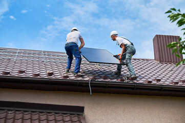 Workers building solar panel system on roof of house. Two men installers in helmets carrying photovoltaic solar module outdoors. Alternative, green and renewable energy generation concept.