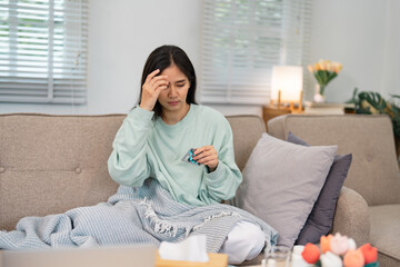 Woman in a mint green sweater sitting at home, rubbing her forehead in distress while holding a...