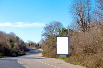 Billboard on the side of the asphalt forest road. White vertical advertising poster on the way....