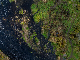 Top-down aerial photo of the Jagala River near Jägala Waterfall in Estonia. On the left the river flows with dark, almost black water speckled with white foam. On the right rugged green landscape.