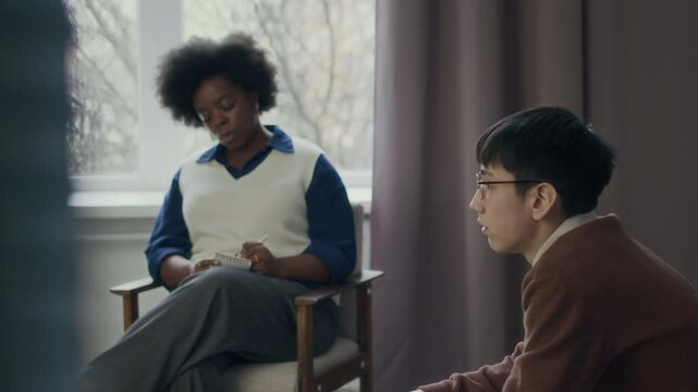 Slowmo of introverted Asian teen boy in eyeglasses sharing worries avoiding eye contact with young African American female counselor during individual therapy session in office