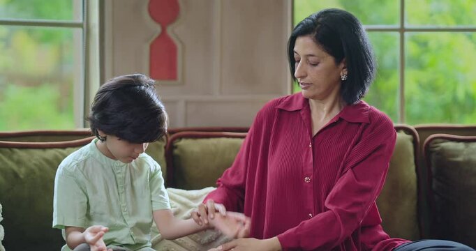 Indian happy relax young adult female lady with cute little son boy kid child teach yoga asana home family sitting on cozy sofa couch talk learn good body health care habit have joy fun enjoy day time