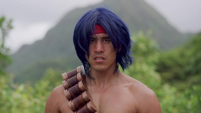 Blue haired, shirtless man with headband and ammunition belt mumbling and glaring at camera while in tropical area