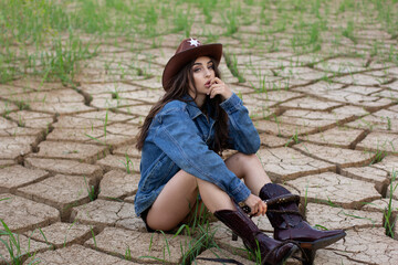 A young woman in a cowboy hat, denim jacket, and boots holding a whip and posing on cracked earth, with a dramatic western-style backdrop.