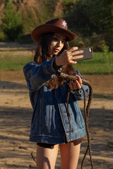 A young woman in a cowboy hat, denim jacket, and boots holding a whip and posing on cracked earth, with a dramatic western-style backdrop.