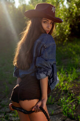 A young woman in a cowboy hat, denim jacket, and boots holding a whip and posing on cracked earth, with a dramatic western-style backdrop.
