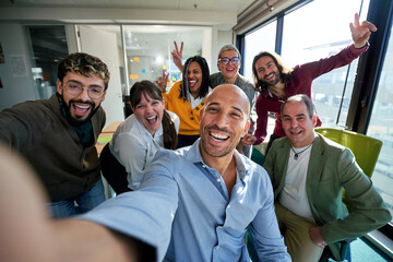 Diverse businesspeople smiling and taking a selfie together in a bright, modern office, showing on social media teamwork and camaraderie between work.