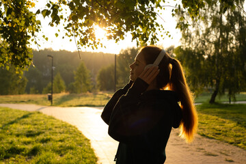 Young woman listening to music and doing breathing exercises in the open air. Red-haired girl meditates while standing against the background of nature.