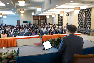 A speaker, viewed from behind, addresses a large audience in a conference hall. The room is filled with rows of attendees seated at tables, listening attentively.