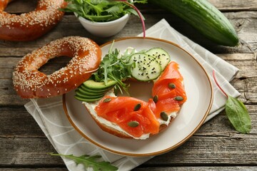Delicious bagel with salmon, cream cheese, cucumber, avocado and pumpkin seeds on wooden table, closeup
