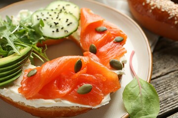 Delicious bagel with salmon, cream cheese, cucumber, avocado and pumpkin seeds on wooden table, closeup